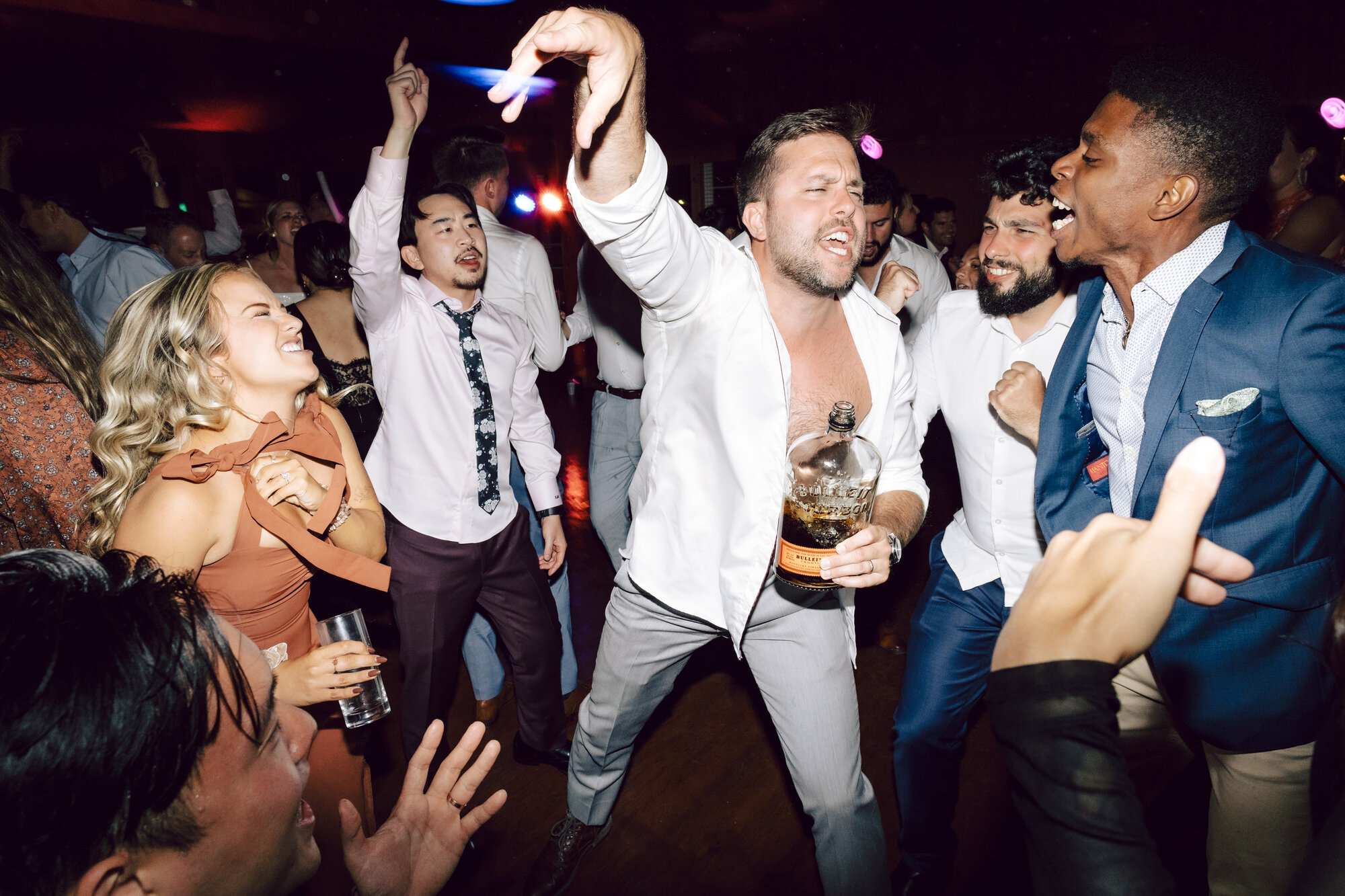 Reception guests singing and dancing as a groomsman with open shirt holds a bottle of bourbon