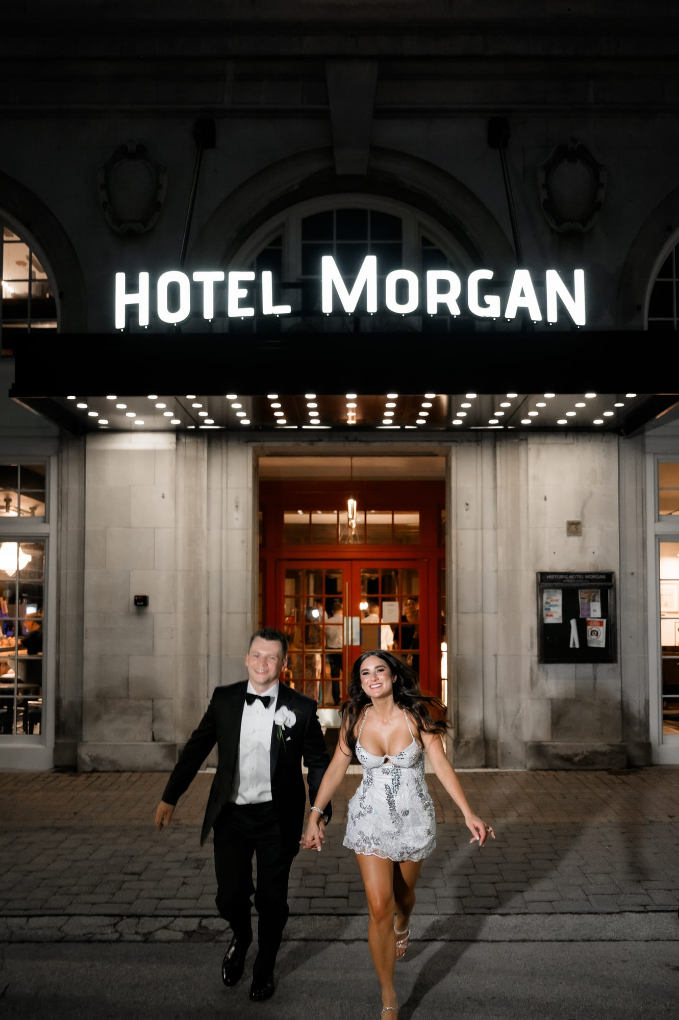 Bride and groom running hand in hand under the glowing Hotel Morgan marquee at night