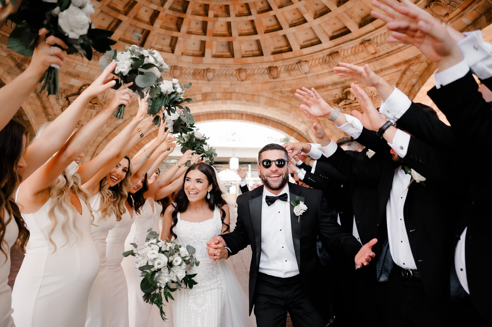 Bride and groom walking through a tunnel of cheering bridal party arms beneath the coffered dome at The Pennsylvanian