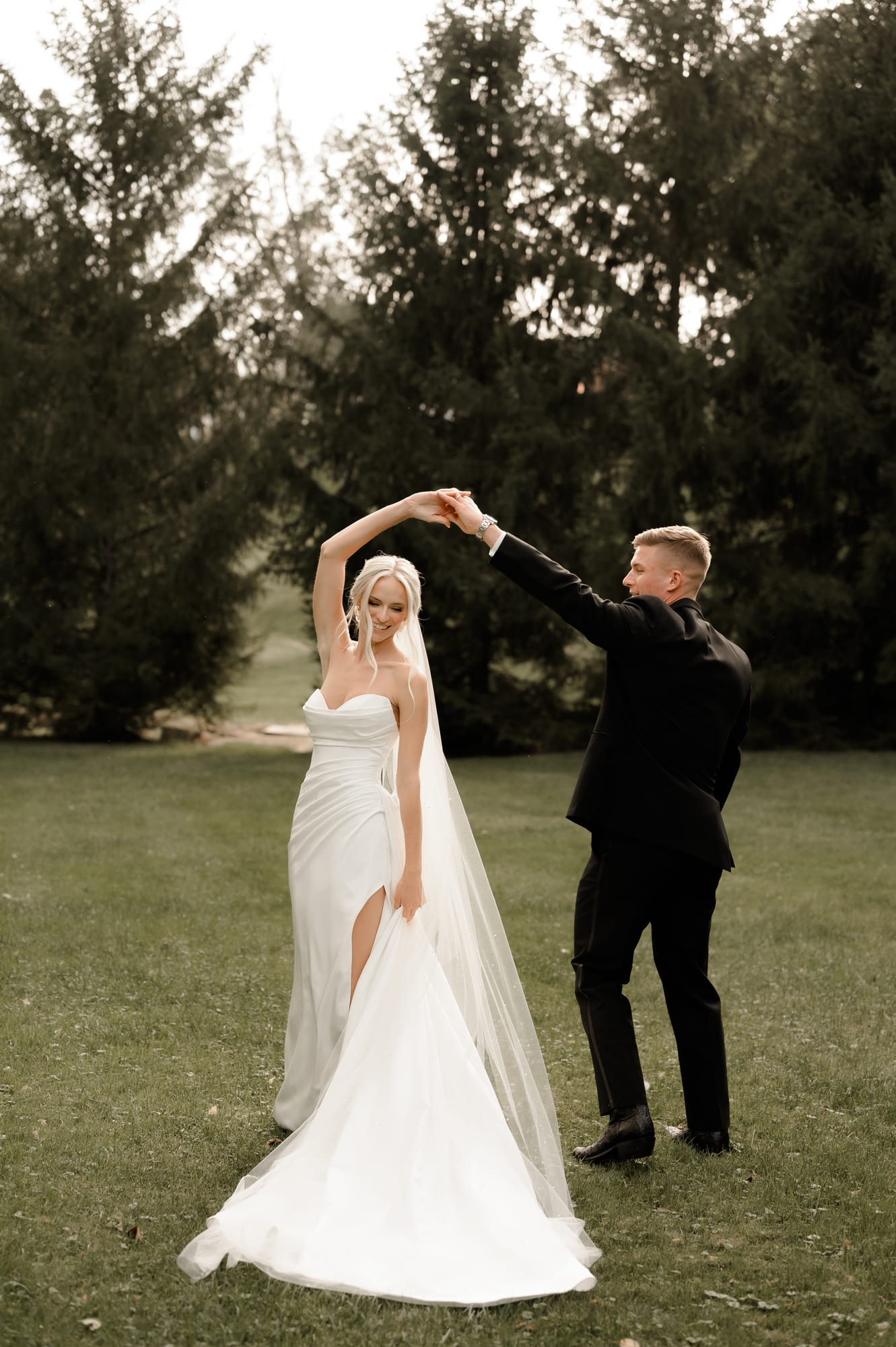 Groom twirling his bride in a wide grass clearing framed by tall pine trees