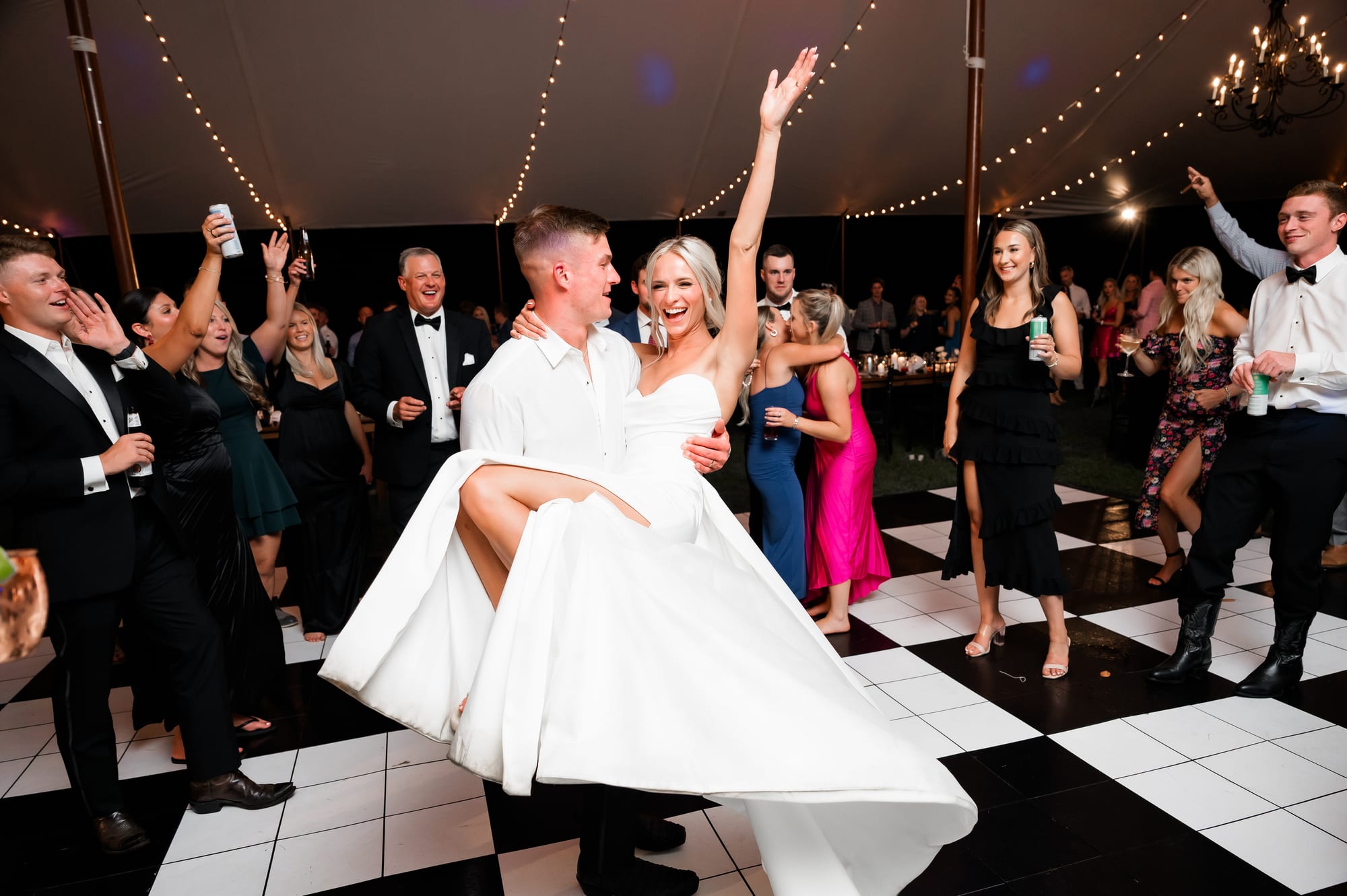 Groom lifting his bride on a checkered tented dance floor with string lights overhead