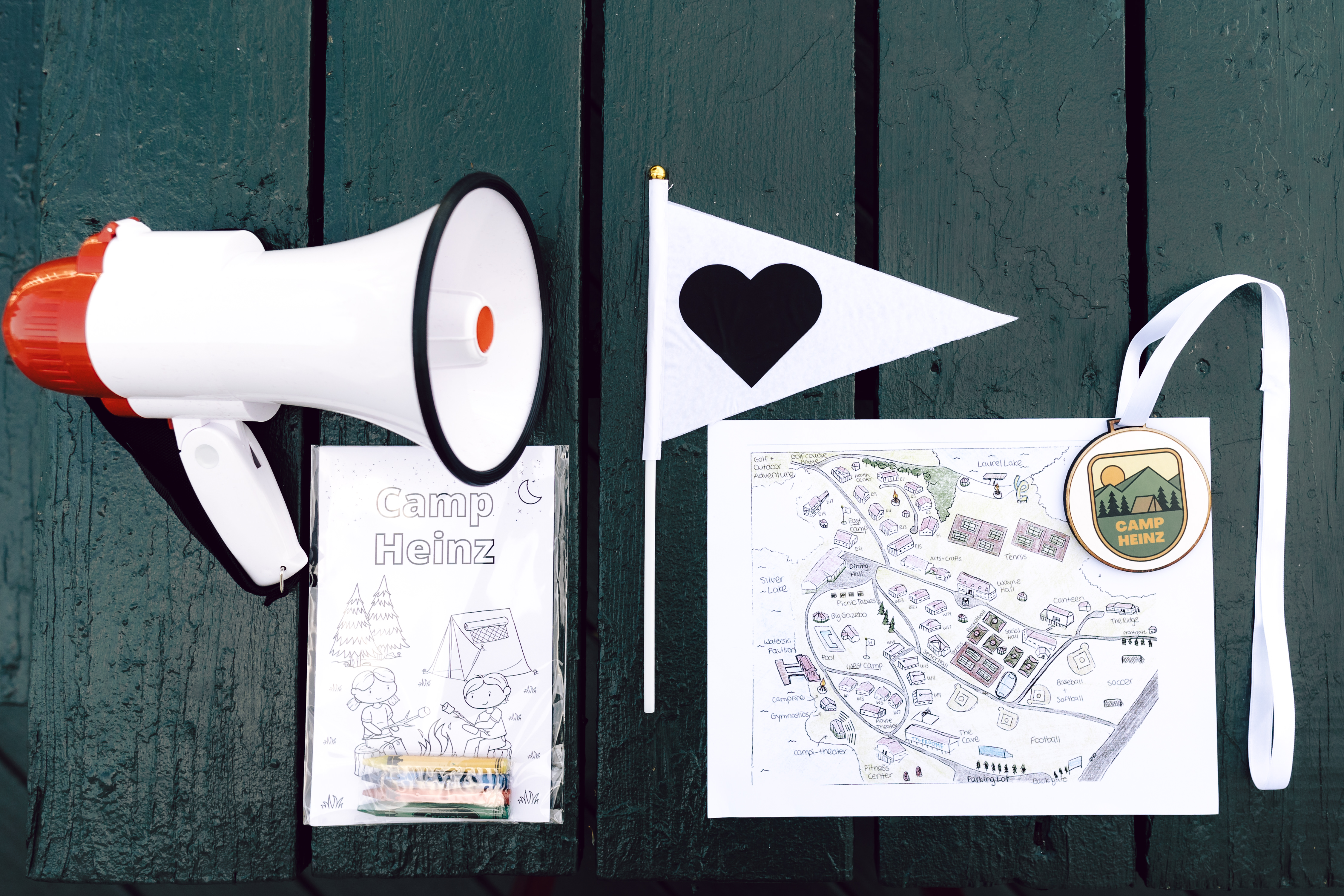 Camp Heinz summer camp themed wedding weekend flat-lay: megaphone, pennant flag with heart, hand-drawn camp map, custom Camp Heinz coloring book and lanyard medallion